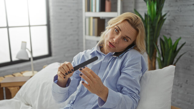 Woman multitasking at home files nails while talking on phone in bedroom showcasing blonde hair and relaxed indoor atmosphere. - Powered by Adobe