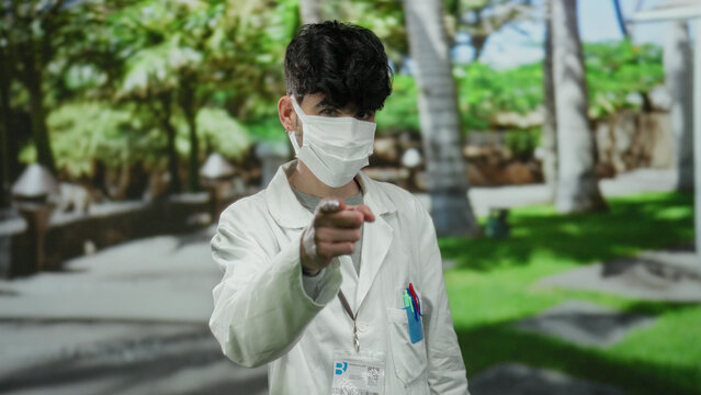 Young man wearing mask in white coat standing in park outdoor with blurred green background during daytime, pointing at face mask with pens in pocket.