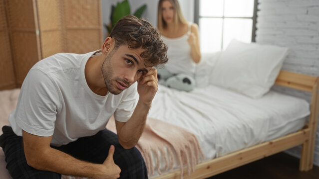 Man and woman in bedroom having conversation with concerned expressions, sitting on bed with neutral decor, implying relationship dynamics and personal reflection indoors.