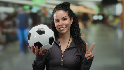 Woman holding soccer ball and making peace sign in an airport terminal, symbolizing sports and...