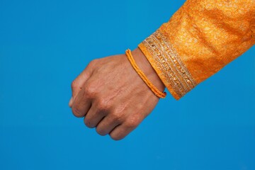 A man's hand with a yellow rakhi on his wrist in a festive outfit in golden yellow tones. On a blue background