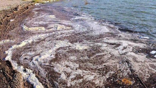 Thick white foam churning over dark, contaminated water, exposing severe ecological damage spreading across polluted shoreline with toxic waste patterns
