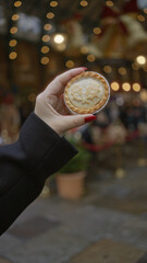 Hand holding festive mince pie in bustling covent garden, london, surrounded by christmas lights...