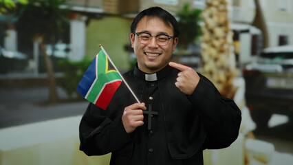 Asian priest holding south african flag smiles on urban street background wearing glasses in...