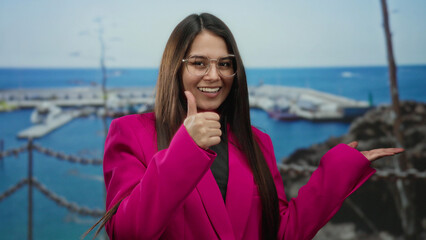 Woman wearing pink jacket pointing and giving thumbs up by the seaside with a marina and blue sea...