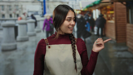 Woman wearing an apron smiles outdoors on a lively street, embodying her role as a waitress in a...