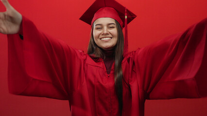 Woman smiling in graduation gown and cap on red background, extending arms in celebration and joy.