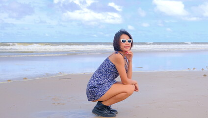 Pretty Asian woman in a floral sundress and black ankle boots, crouching on a beach against a serene ocean backdrop