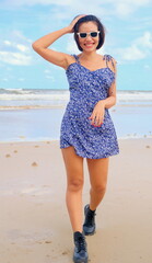 Confident young Asian woman walks toward the camera on the beach in a stylish blue floral dress, paired with bold black boots