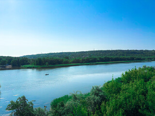 A wide tranquil river flows calmly under a clear blue sky, surrounded by lush green trees and forest-covered hills. 