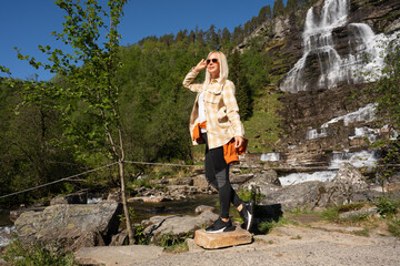 Naklejka premium A young female traveler is enjoying the beautiful sunny weather and a stunning waterfall view during a hiking trip in the mountains.