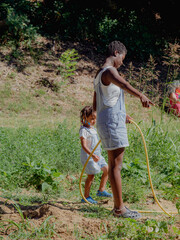 African American mother and daughter with black hair collaborating, watering vegetable garden...