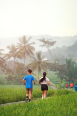 Obraz premium Rear View Asian Couple Jogging Together on Rural Path Surrounded by Green Fields in the Morning