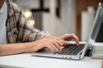 Close up of a man in scotch plaid shirt typing on laptop with both hands sits at white table in cafe