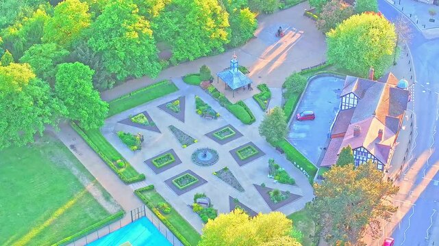 Aerial view shows geometric formal garden with central fountain, pavilion, and Tudor-style house in Matlock Sparrow Park, Derbyshire, England, surrounded by greenery and paths, drone shot
