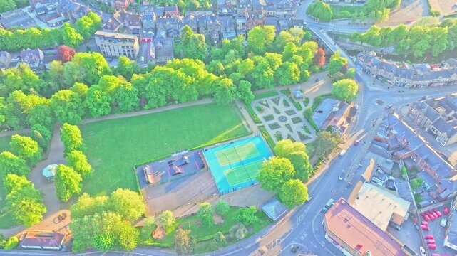 Aerial scene reveals Matlock&rsquo;s Sparrow Park in Derbyshire, with green lawns, tennis courts, geometric gardens, tree canopies, pathways, and adjacent town streets, showcasing urban green space