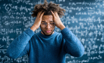 Frustrated student in blue sweater holding head in front of chalkboard covered with complex math equations.