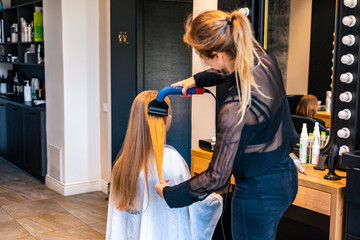 Female hairstylist drying and straightening long blonde client hair with comb and hairdryer in...