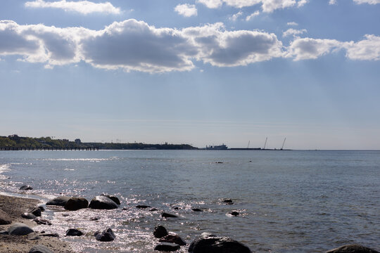 Rocky seashore with calm waves and distant port under partly cloudy sky. Industrial coastline view with ship and cranes.