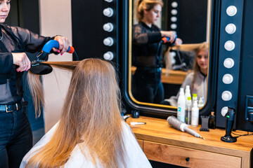 Female hairstylist drying and straightening long blonde client hair with comb and hairdryer in...
