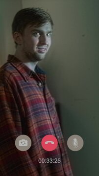 Vertical handheld shot of cheerful girl chatting on video call while creepy man standing by window then turning and grinning at camera under lightning flashes in living room