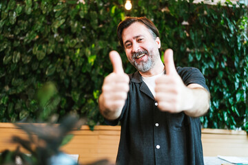 Happy middle-aged cafe owner giving two thumbs up in front of a lush green wall. He smiles confidently, celebrating business success and customer satisfaction in a cozy, eco-friendly setting.