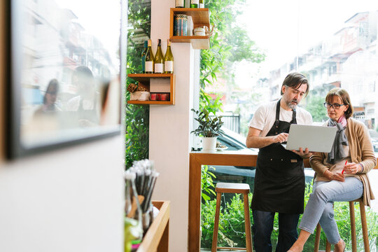 Cafe owner and financial advisor discussing business strategy using a laptop in a modern coffee shop. Bright setting with plants and wine bottles, symbolizing collaboration and entrepreneurship.