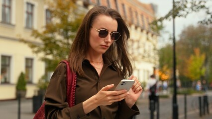 Obraz premium Confident stylish Caucasian woman walking in city holding mobile phone walk at street using smartphone online internet app chatting female girl fashionable wearing sunglasses typing cellphone media