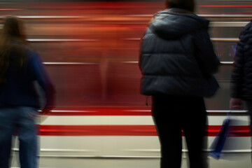 Blurred figures of people standing on a subway platform with a red and white train speeding past,...