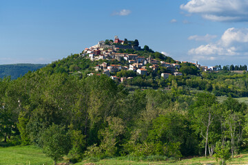 A picturesque hilltop town, possibly in Istria, Croatia, with charming houses and a church, surrounded by lush green trees under a clear blue sky.