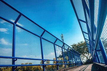 A low angle view of a vibrant blue pedestrian bridge featuring strong mesh barriers leading towards...