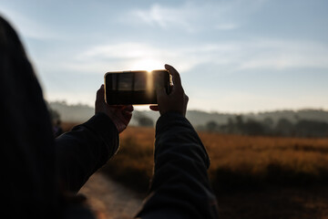 person is seen holding a smartphone, capturing a sunrise or sunset in a wide field.