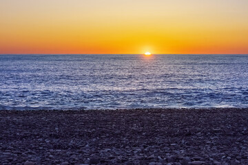 Sunset over darkness sea horizon with pebble beach. Sun is casting warm hues of orange and purple...