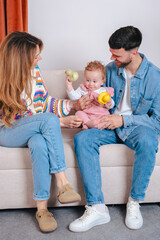Smiling parents on sofa entertain their baby daughter in pink overalls with yellow duck and green...