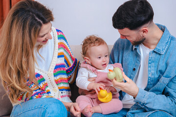 Smiling parents on living room sofa entertain their baby daughter in pink overalls with yellow duck and green turtle toys, capturing playful family bonding and early childhood development indoors. 