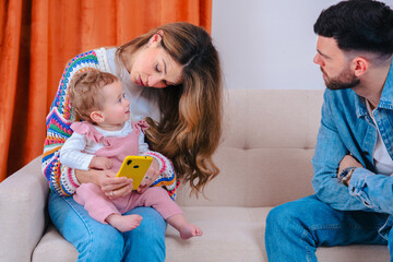 Tense family moment where a father looks upset while the mother shows cartoons to their baby on a...