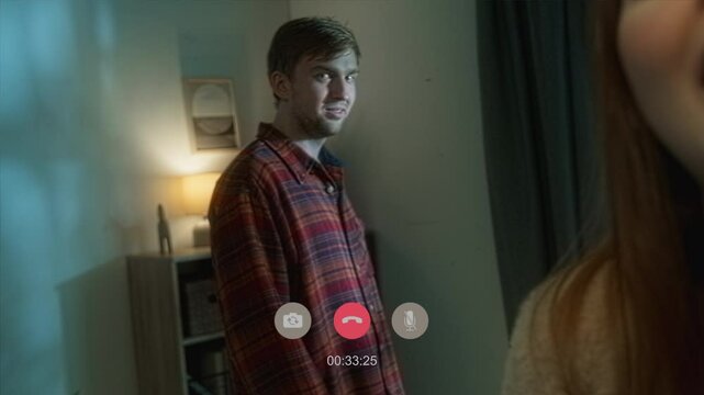 Handheld shot of cheerful woman talking on video call while spooky man standing by window staring at lightning then turning and grinning at camera in living room