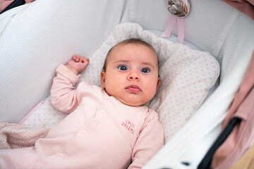 A baby with big eyes lies in a cozy stroller, dressed in a soft pink outfit. The infant appears...