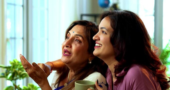 Mother daughter having morning coffee together near window, Indian asian old mom in saree and young daughter standing in modern home enjoying hot drink while watching nature view and bonding happily