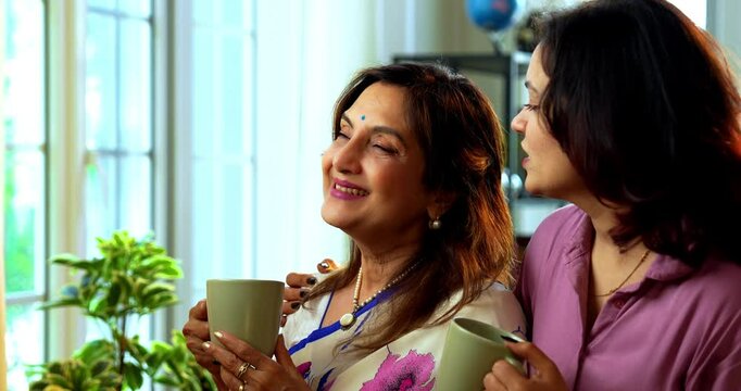 Mother daughter having morning coffee together near window, Indian asian old mom in saree and young daughter standing in modern home enjoying hot drink while watching nature view and bonding happily