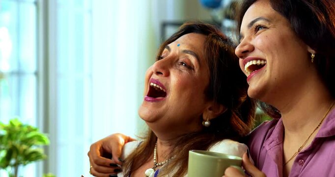 Mother daughter having morning coffee together near window, Indian asian old mom in saree and young daughter standing in modern home enjoying hot drink while watching nature view and bonding happily
