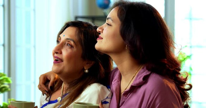 Mother daughter having morning coffee together near window, Indian asian old mom in saree and young daughter standing in modern home enjoying hot drink while watching nature view and bonding happily