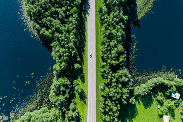 Aerial view of electric car driving asphalt road between blue water lake and green woods in Finland