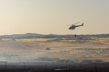 A yellow firefighting helicopter flies through thick smoke over charred terrain during a scorching...