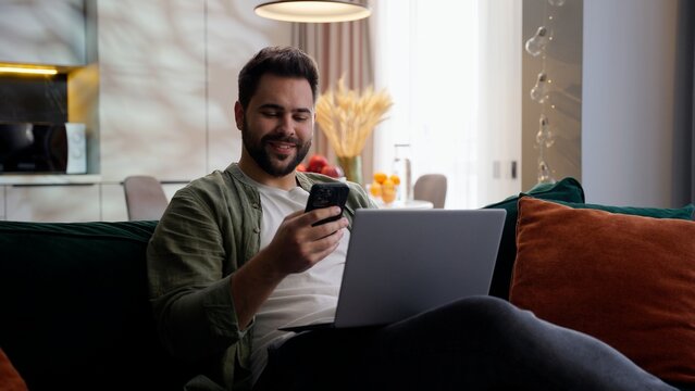 Happy joyful Caucasian business man male guy entrepreneur businessman freelancer client sitting sofa work laptop computer hold mobile phone showing smartphone green screen chroma key mock up at home - Powered by Adobe