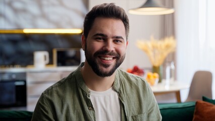 Obraz premium Cheerful happy European Caucasian young adult bearded man male guy homeowner laughing smiling joyful model businessman look distance watching TV looking at camera laugh indoors living room home rent