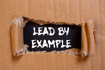 Lead by example message written on ripped torn cardboard paper on black background. Conceptual lead...