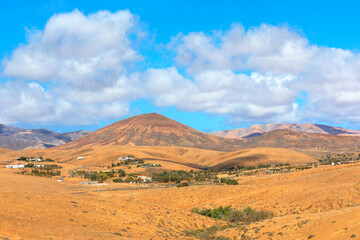 Scenic Canary Islands landscape with rolling golden hills under blue sky dotted with fluffy clouds. Scattered white houses nestle in valleys, surrounded by arid terrain and distant rugged mountains