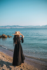 Woman in black dress and hat looking at sea on greek beach