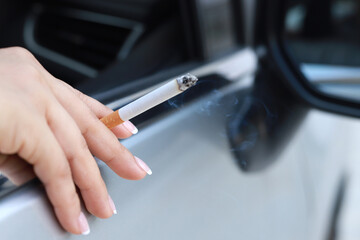 Hand holding burning cigarette while gripping steering wheel, with smoke filling car.